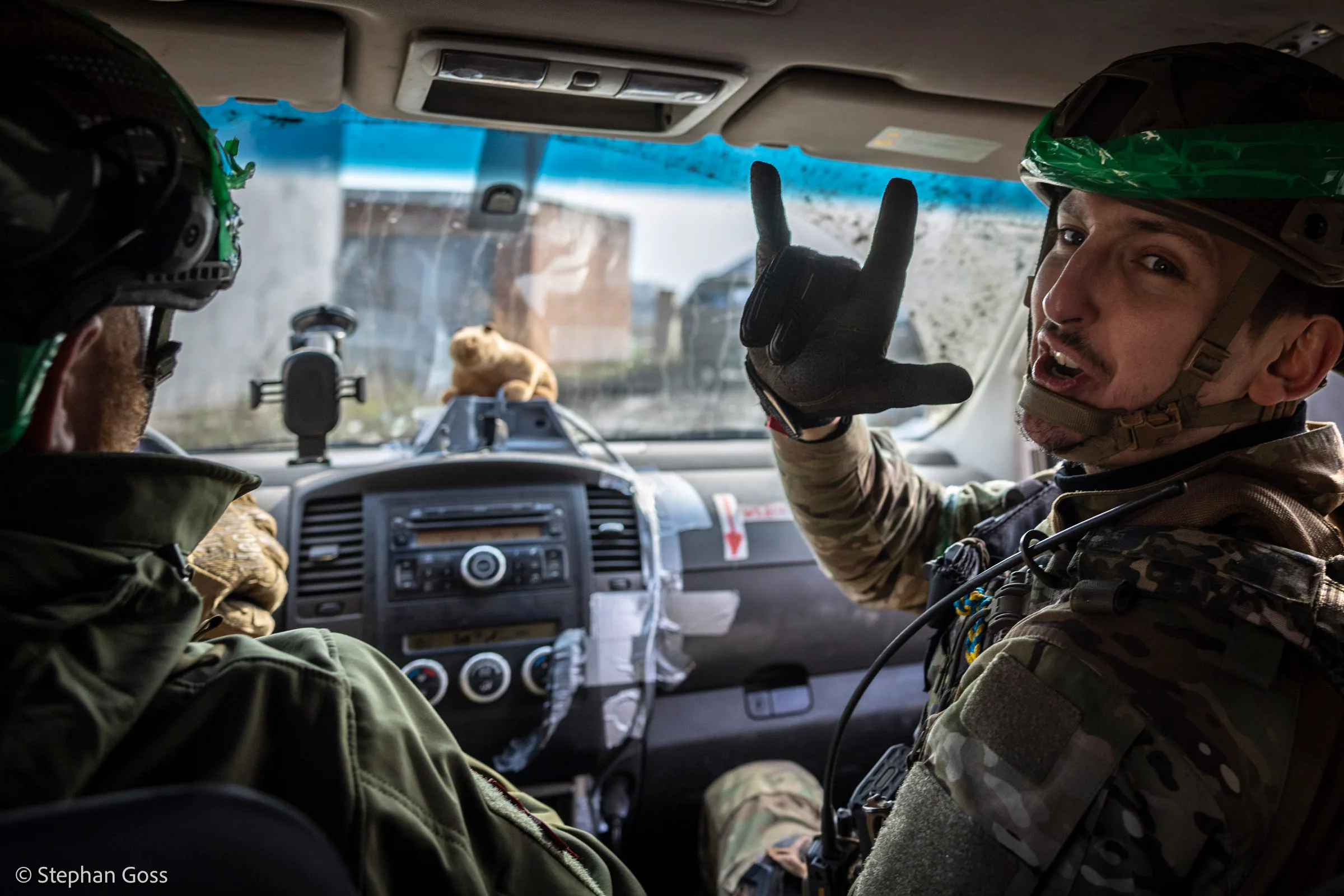 Two soldiers in the cab of a vehicle; the one in the passenger seat turns to the camera with his hand raised, a soft toy sitting on the dashboard behind a cracked windscreen.