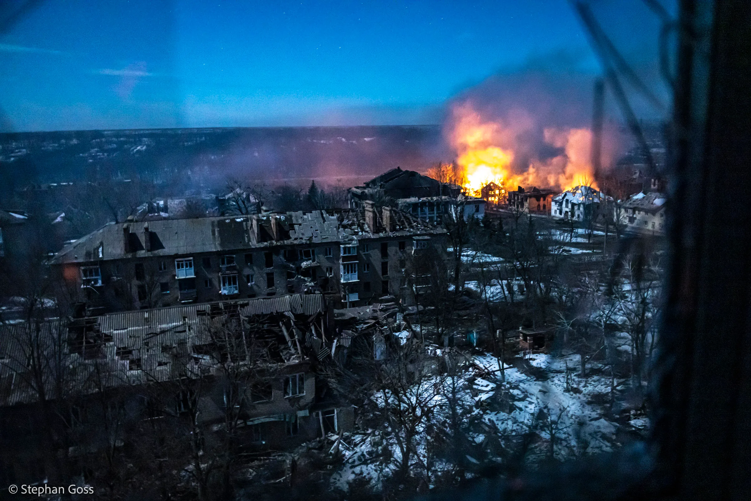 Seen from a high window at dusk, a town of roofless, burnt-out apartment blocks stretches away under a deep blue sky, an orange fire burning in the middle distance.