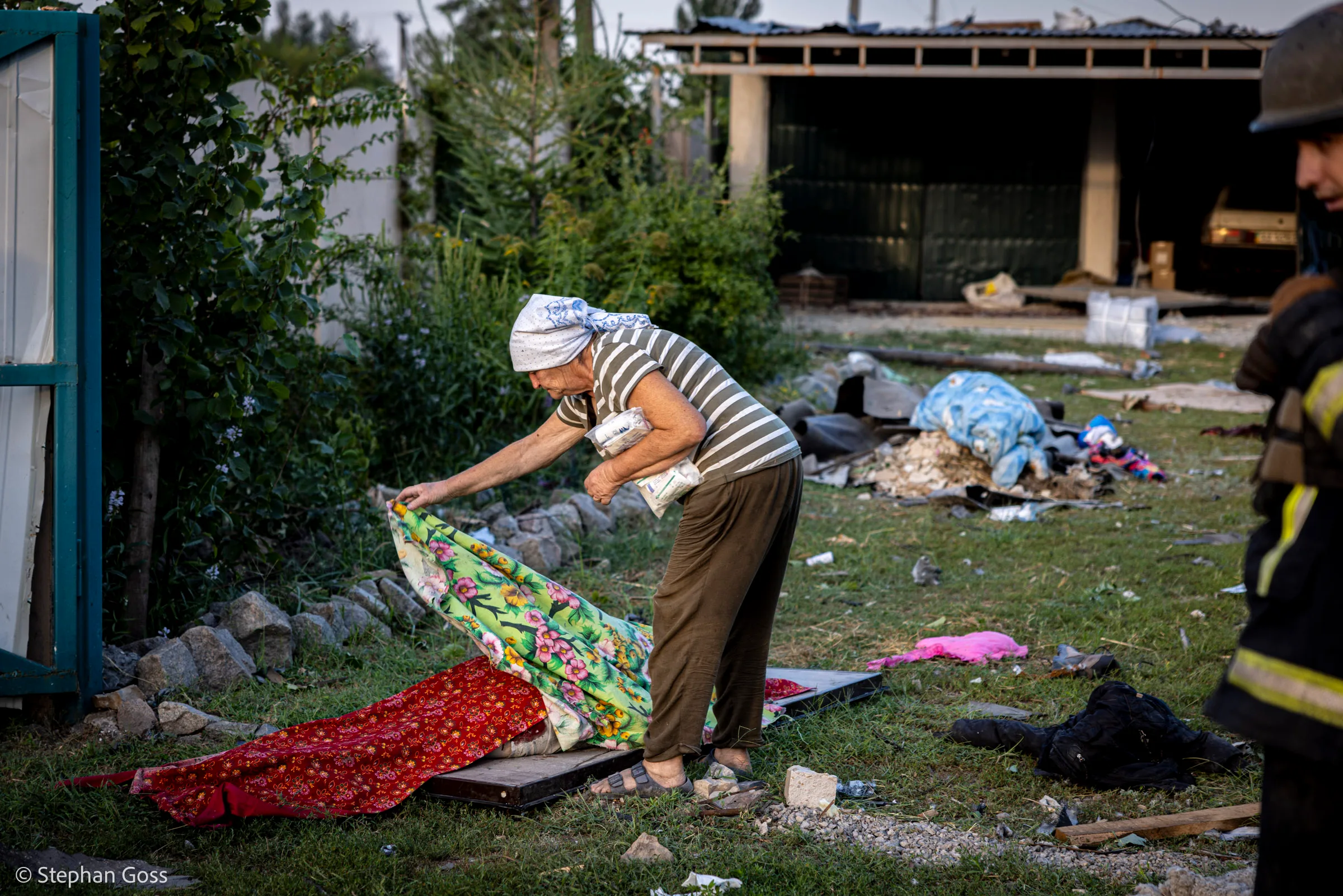An elderly woman in a headscarf lifts a floral quilt from the grass outside a damaged house, bedding and debris scattered around her and a firefighter at the edge of the frame.